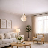 A bright, airy living room features a white sofa, round wooden coffee table with flowers, wicker chair, potted plant, framed wall art, large window with sheer curtains, and the Virello 1 Light Small Raindrop Alabaster Single Pendant Light in Soft White.
