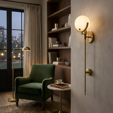 A cozy reading nook with a green armchair, a round marble side table, brushed brass floor lamp, built-in wooden shelves, and the Solara 1 Light Large Sphere Stem Alabaster Wall Light in soft white. Large windows showcase a city view.