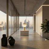 A modern, minimalistic room with large glass windows showcases a city skyline at sunset. The Dipping Table Lamp - Gold, with its colourful glass globe and brushed brass base, sits on a stone pedestal beside a potted plant.