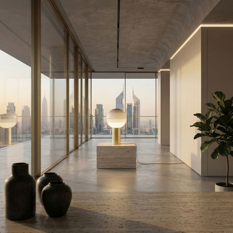 A modern, minimalistic room with large glass windows showcases a city skyline at sunset. The Dipping Table Lamp - Gold, with its colourful glass globe and brushed brass base, sits on a stone pedestal beside a potted plant.