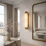 A modern, elegant living room with tall windows, beige curtains, a large wall mirror, a marble table with pink flowers, and the Orivelle LED Small Vertical Alabaster Wall Light in soft white and brushed bronze emits warm light.