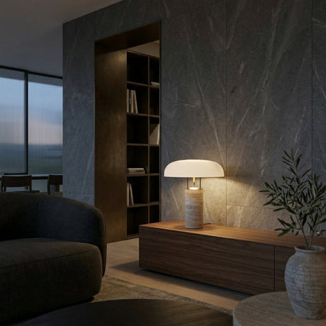 A modern living room with dark stone walls, a wooden low cabinet, the Caldrin 1 Light Stone Column Table Lamp in grey & white, a gray sofa, a green plant, bookshelves in the background, and evening light filtering through large windows.