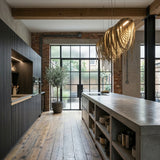 Modern industrial kitchen with exposed brick walls, large factory-style windows, a concrete island with open shelving, dark cabinetry, wood floors, a potted plant, and the elegant Symphonia 10 Light Metal Chain Veil Suspension in gold as a statement chandelier.