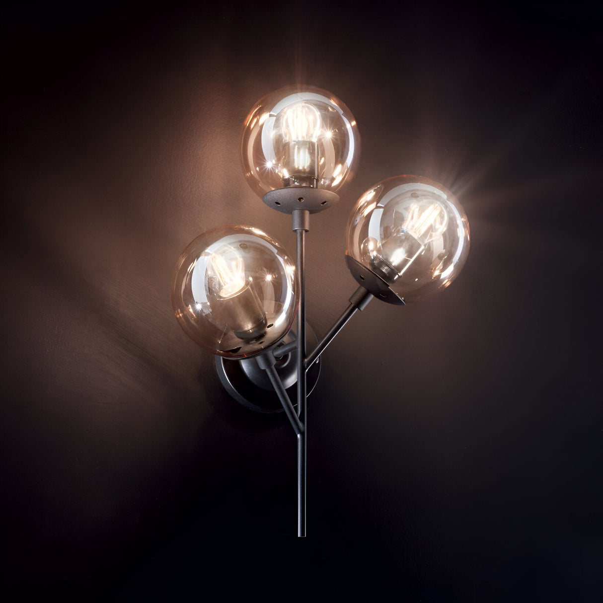 The Novara 3 Light Wall Light - Black showcases a refined black metal frame with three luminous glass globes, complemented by amber blown glass diffusers. Mounted on a dark wall, it emits a cozy and welcoming light.