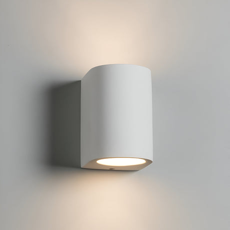 A contemporary, minimalist Curved Up And Down Wall Light with a cylindrical design is mounted on a light gray wall. Its gypsum construction channels ambient lighting both upwards and downwards, resulting in a gentle illumination.