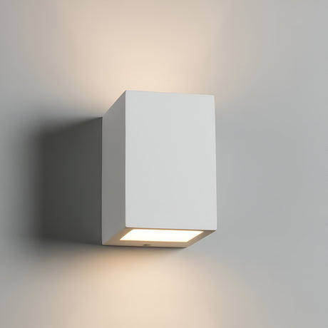 The Cuboid Up And Down Wall Light, a minimalist rectangular plaster fixture, is mounted on a light-colored wall. The dimmable light illuminates the space with a soft glow above and below, providing ambient lighting and creating a subtle shadow effect.