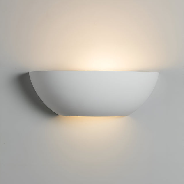 The Up And Down Plaster Wall Light is a contemporary, white wall sconce featuring a half-circle design that provides an elegant touch. Installed on a light-colored wall, it casts a warm, soft glow upwards and downwards, enhancing the cozy ambiance with its soothing ambient lighting.