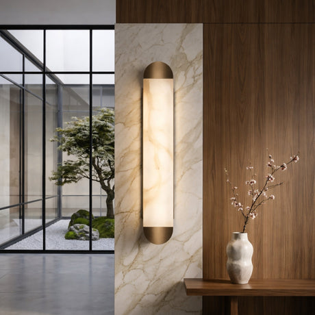 A modern interior with a marble and wood wall, the Orivelle LED Medium Vertical Alabaster Wall Light in soft white & brushed bronze, a vase with branches on a wooden console, and a view of a minimalist courtyard with tree and rocks.