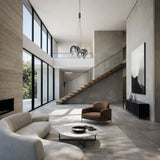 A modern, minimalist living room with large windows, neutral-toned furniture, a curved sofa, circular coffee table, brown armchair, floating wooden staircase, abstract wall art, and the Diverso LED Pendant Light - Chrome as a contemporary lighting fixture.