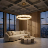 A modern living room with a curved beige sofa, round coffee table with pottery, large windows revealing a sunset city skyline, wooden panel walls, and the Orion 36 Light Ring Alabaster Chandelier in Soft White & Brushed Brass on a coffered ceiling.