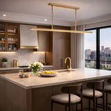 A modern kitchen with light wood cabinets, marble countertops, a large island with bar stools, brushed brass fixtures, fresh tulips in a vase, a bowl of lemons, and an Elvaris LED Linear Alabaster Suspension Light illuminating the city skyline at sunset.