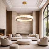 A modern, elegant living room with cream sofas and chairs, a round coffee table, floor-to-ceiling window, light beige walls, and the Orion 36 Light Ring Alabaster Chandelier in soft white & brushed brass hanging from the high ceiling.