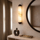 The Orivelle LED Small Vertical Alabaster Wall Light in Soft White & Brushed Bronze is mounted on a textured wall beside a round mirror. Below, a sculptural decor piece and tray with wooden beads rest on a light console table.