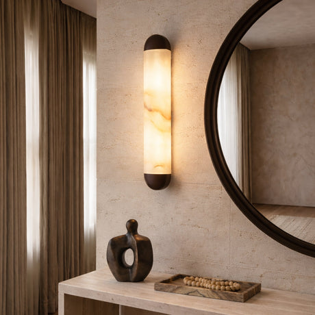 The Orivelle LED Small Vertical Alabaster Wall Light in Soft White & Brushed Bronze is mounted on a textured wall beside a round mirror. Below, a sculptural decor piece and tray with wooden beads rest on a light console table.