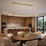 A modern kitchen and dining area with wooden cabinets, a marble island with wicker barstools, round wooden dining table, cushioned chairs, and the Elvaris LED Alabaster Suspension Light in brushed brass. Glass doors open to a patio with seating and greenery.