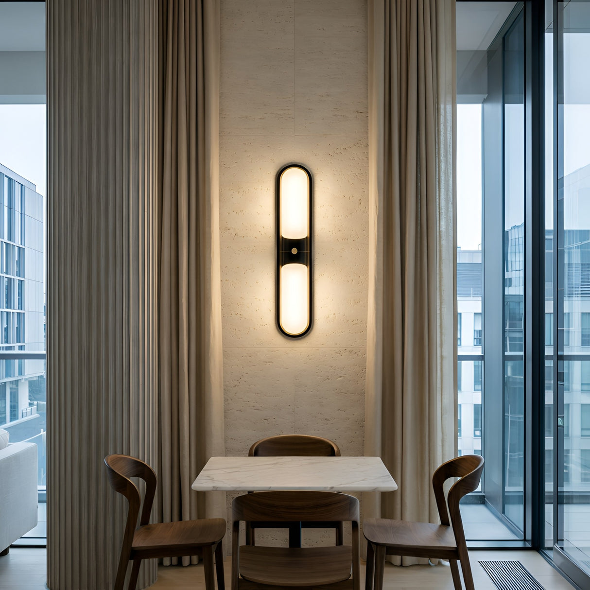 A modern dining area with a small marble table, three wooden chairs, floor-to-ceiling curtains, and the Lunaris LED Capsule Alabaster Wall Light in soft white & black. Large glass windows reveal contemporary buildings outside.