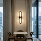 A modern dining area with a small marble table, three wooden chairs, floor-to-ceiling curtains, and the Lunaris LED Capsule Alabaster Wall Light in soft white & black. Large glass windows reveal contemporary buildings outside.