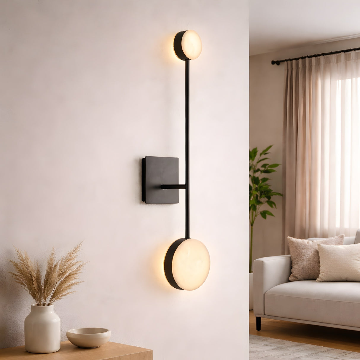 The Marvello LED Twin Sphere Alabaster Wall Light in soft white and matte black is mounted on a white wall in a minimalist living room with neutral decor, a beige sofa, and large windows draped with sheer curtains.
