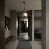 A modern hallway with gray walls and floors, minimal walnut brown shelves, a built-in bench, a Lunaro 1 Light Small Travertine Single Pendant Light in Walnut Brown & Light Beige, and a textured rug, leading to a sunlit room.