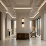 A modern living room with high coffered ceilings, large windows, beige walls, a marble fireplace, two armchairs, a wooden sideboard, and a central partition featuring the Lunaris LED Capsule Alabaster Wall Light in soft white & black.