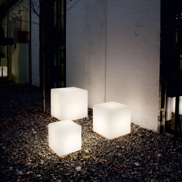 Inspired by the minimalist design of the Spectra Outdoor Pedestal Light - White, 30cm, three glowing cube-shaped lights are arranged on a pebble-covered outdoor area against a white brick wall. They emit a soft, warm glow that subtly enhances the space with outdoor lighting.
