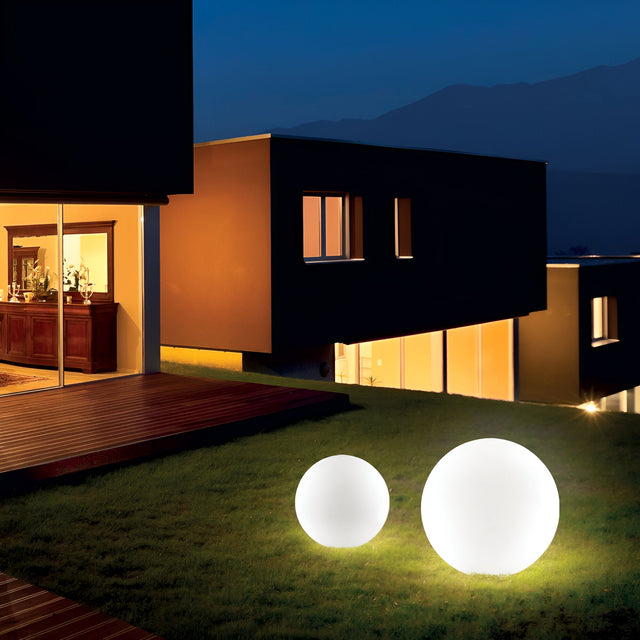 On a quiet night, a modern house with expansive illuminated windows stands prominently. In the foreground on a well-kept lawn, two Lucidra Outdoor Pedestal Lights in white, each featuring 40cm opal plastic diffusers, glow softly and enhance the tranquility of the dark landscape around them.