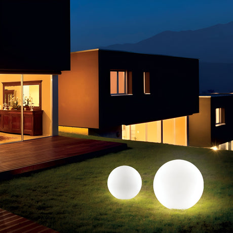 A modern house at night showcases large windows softly lit from within. The garden is illuminated by two Lucidra Outdoor Pedestal Lights in white, each 30cm in diameter, featuring an aluminum suspension base that highlights the garden lighting. The surrounding landscape remains dimly visible under a dark blue sky.