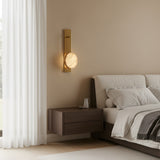 A modern bedroom with a beige wall, a bed with white and gray bedding, a floating wooden nightstand, and the Marvello LED Sculpted Disc Alabaster Wall Light in soft white & brushed brass glowing warmly next to sheer white curtains.