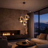 A modern living room with a marble coffee table, beige sofas, a lit fireplace, abstract sculpture, city-view windows at dusk, and three Diverso LED Pendant Lights in gold hanging from the ceiling.