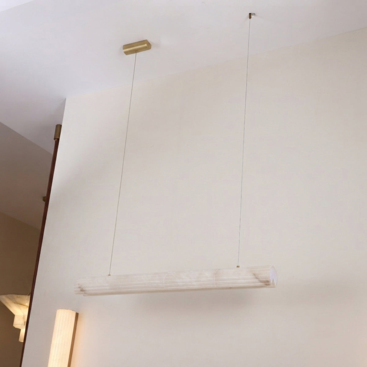 Zenvia LED Alabaster Linear Chandelier in Soft White, a horizontal alabaster cylinder pendant on twin brass cables from a slim ceiling fixing, shown against a plain wall.