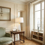 A cozy, elegant living room with cream walls, a large window with light curtains, a green armchair, wooden side table featuring the Arvello 1 Light Slim Tapered Marble Table Lamp in Brass & White, built-in shelves with books and decor, and a wall mirror.