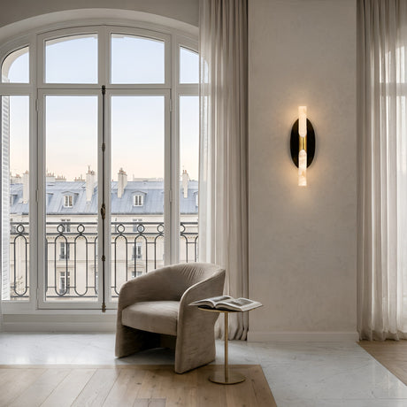 A modern, minimalist room features a curved beige armchair beside a table with an open magazine. Large arched windows show Parisian rooftops, while the Virexa LED Oval Backplate Alabaster Wall Light glows on a textured wall. Light curtains frame the scene.