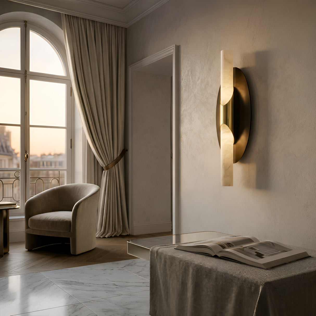 A softly lit, elegant room features a curved armchair by a tall window with cream curtains, marble floor, and the Virexa LED Oval Backplate Alabaster Wall Light in Soft White & Black. An open book rests on a fabric-covered bench under natural light.