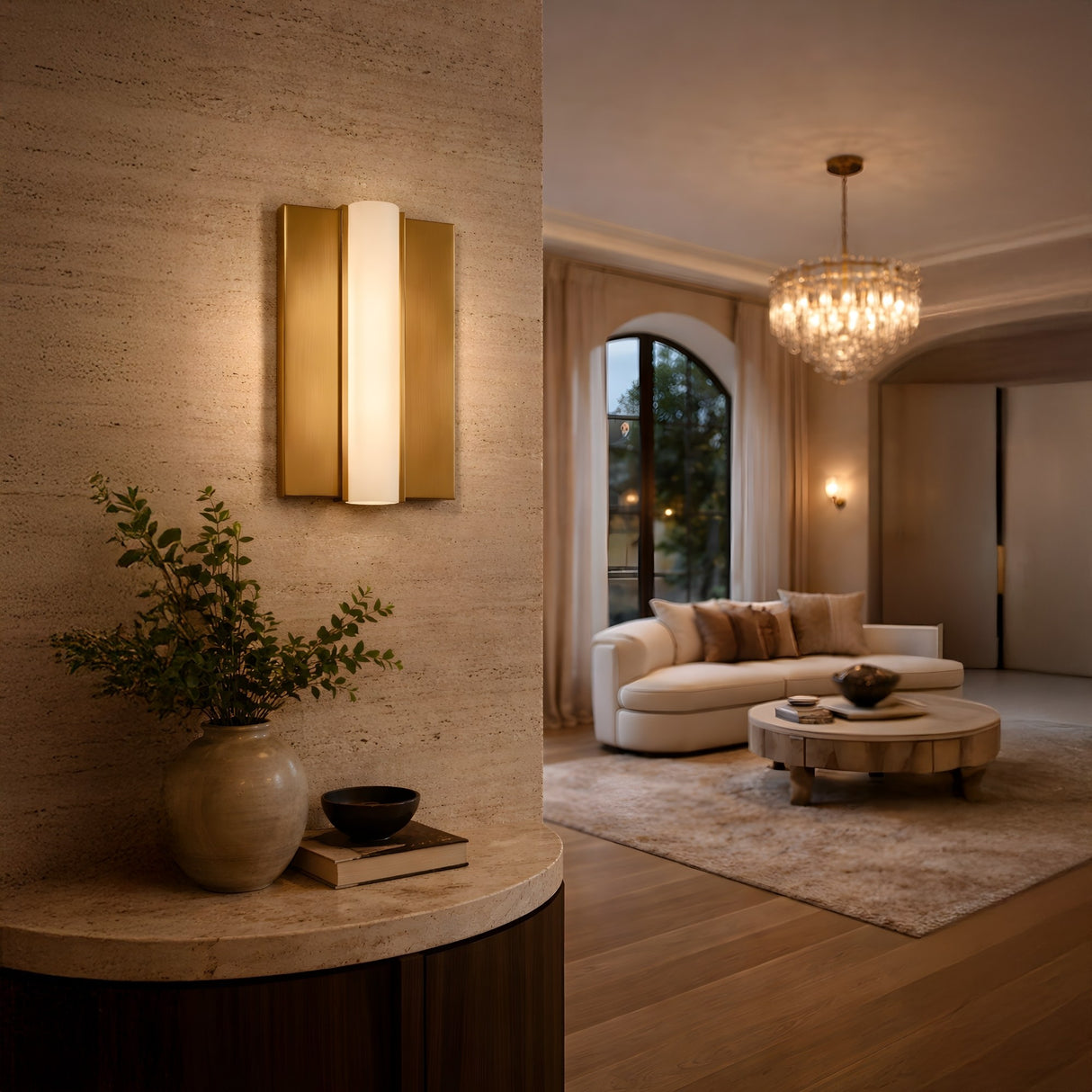 A modern, warmly lit living room features the Elvaris LED Small Vertical Alabaster Wall Light in brushed brass & soft white, a vase with greenery on a console, a cozy white curved sofa, round coffee table, beige rug, and chandelier.