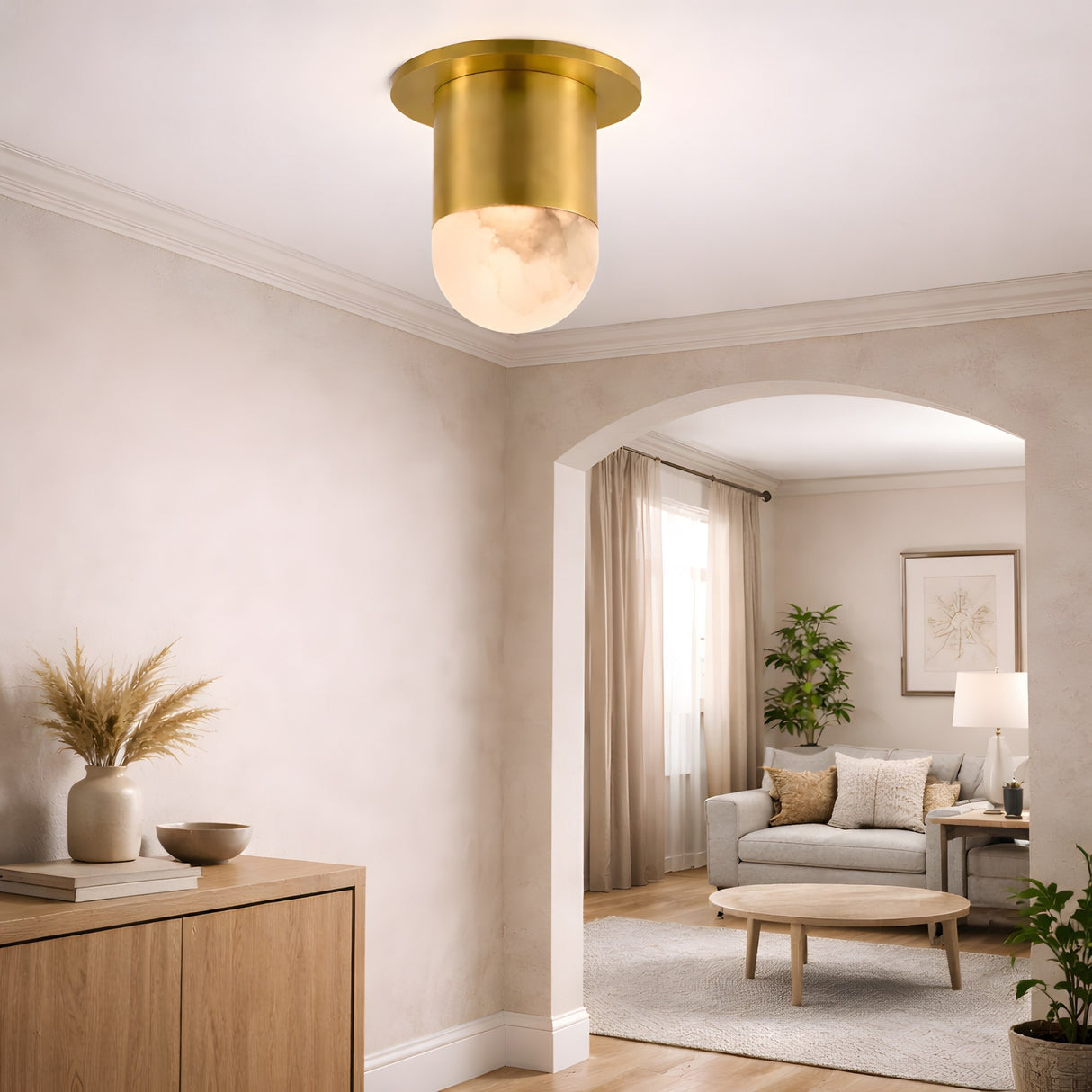 A modern living room with neutral tones showcases the Elara LED Mini Drop Alabaster Semi-Flush Ceiling Light in Soft White & Brushed Brass, an archway, beige walls, a wood sideboard with decor, and a cozy sofa with a round coffee table.