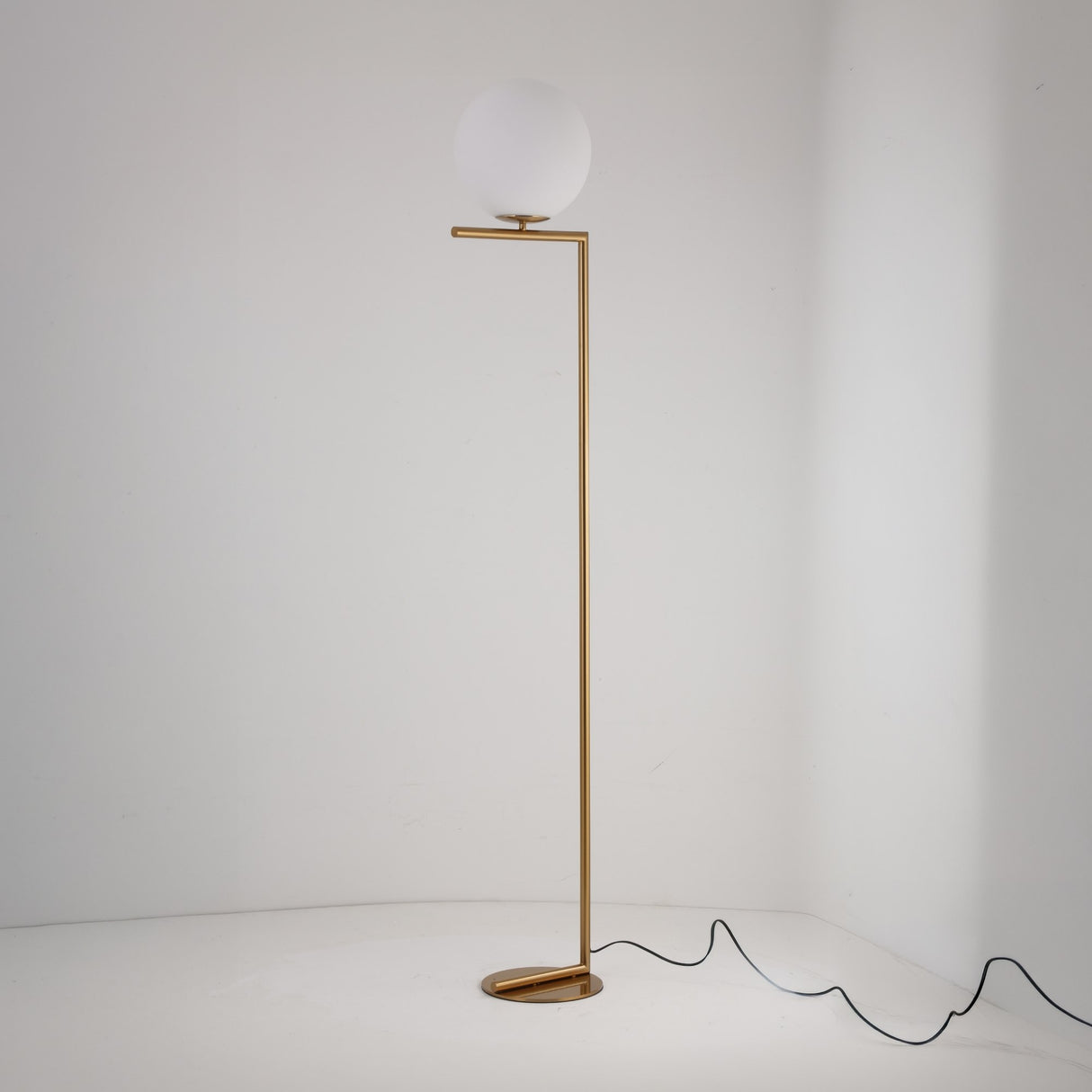 The Vetra 1 Light Opal Glass Floor Lamp in gold features a slim metal stand, horizontal arm, and round white globe shade. Its minimalist design provides soft lighting against a white wall, with a black cord neatly trailing on the floor.
