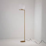 The Vetra 1 Light Opal Glass Floor Lamp in gold features a slim metal stand, horizontal arm, and round white globe shade. Its minimalist design provides soft lighting against a white wall, with a black cord neatly trailing on the floor.