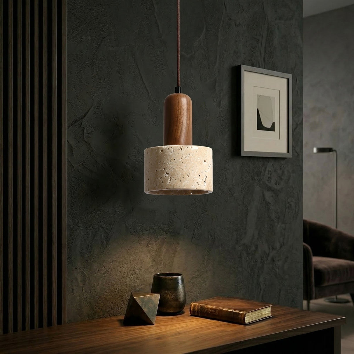 The Lunaro 1 Light Large Travertine Single Pendant Light in Walnut Brown & Light Beige hangs above a wooden desk with a book, ceramic cup, and geometric decor. Dark textured walls and minimalist artwork complete the room’s look.