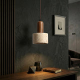 The Lunaro 1 Light Large Travertine Single Pendant Light in Walnut Brown & Light Beige hangs above a wooden desk with a book, ceramic cup, and geometric decor. Dark textured walls and minimalist artwork complete the room’s look.