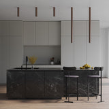 A modern kitchen showcases a black marble island with two black bar stools. The minimalist cabinets are light gray, complemented by a countertop featuring a black sink and a bouquet of yellow flowers. Pulcra LED Tube Ceiling Lights in copper hang above, enhancing energy efficiency, while a bowl of lemons rests on the counter.