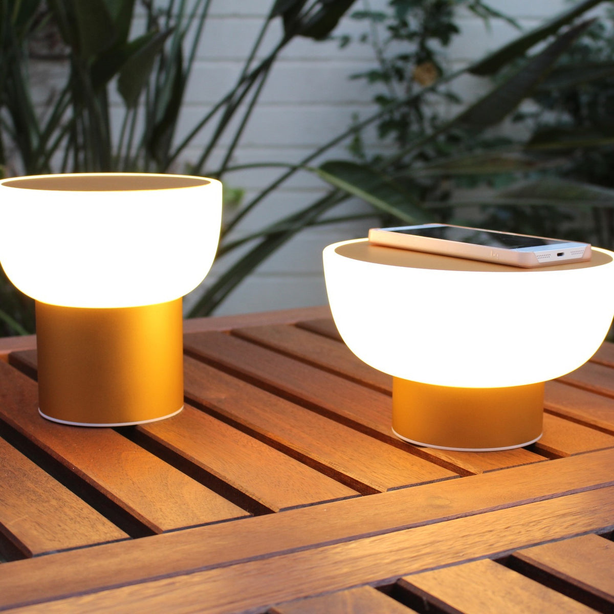 Two Patio rechargeable LED table lamps with white bowl-shaped shades on gold cylinder bases, glowing on a wooden outdoor table beside a smartphone.