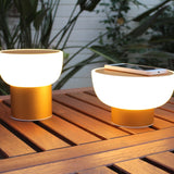 Two Patio rechargeable LED table lamps with white bowl-shaped shades on gold cylinder bases, glowing on a wooden outdoor table beside a smartphone.
