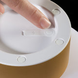 Close-up of the Patio LED rechargeable table lamp's white underside, showing a touch power button, charging icon, and rubber feet above a gold base, with a finger demonstrating the control.