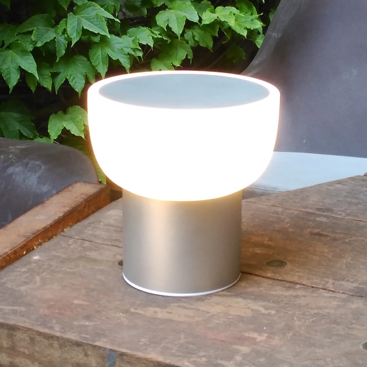 Patio LED rechargeable outdoor table lamp in silver, with a rounded opal shade on a slim cylindrical base, glowing warm white, shown outdoors on a wooden surface.