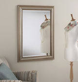 An elegant room decor showcases a mannequin with a floral scarf and pearl necklace next to the Verity mirror, which features a large rectangular silver frame. A striped armchair with a blue pillow and draped pearls peeks in from the left.