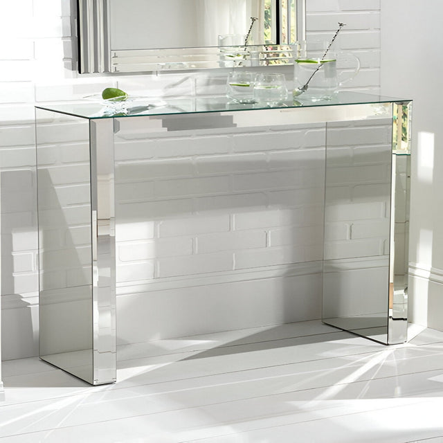 The Atrium Mirrored Console Table - Silver stands against a white brick wall, topped with a glass pitcher, lime-adorned glasses, and a metal straw. Sunlight casts sleek shadows on the white floor, creating a chic modern vibe.