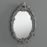 The Mirage Oval Wall Mirror Silver (64x46cm) features a floral-patterned frame, bringing decorative depth and charm to any plain gray wall.