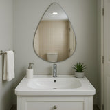 A modern bathroom features a white sink and cabinet, silver faucet, soap dispenser, and plant. An Oculus Droplet Wall Mirror Clear - 90x60cm with a teardrop shape reflects the toilet and beige tiles. A towel hangs on a rack to the left.