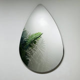 The Oculus Droplet Wall Mirror Clear (90x60cm), with its unique teardrop shape, is mounted on a white wall and reflects green fern leaves along with their shadows.