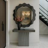 A modern entryway with the Solstice Circle Wall Mirror - Grey, boasting wavy edges above a sleek, vertically lined console table topped with a dark vase of pink and peach flowers; a staircase appears on the right.